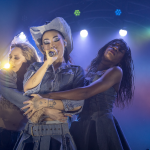 A photo of Rina Sawayama being embraced by backup dancers Shola B Riley (left) & Summer Jay Jones (right) with purple, blue and yellow light.